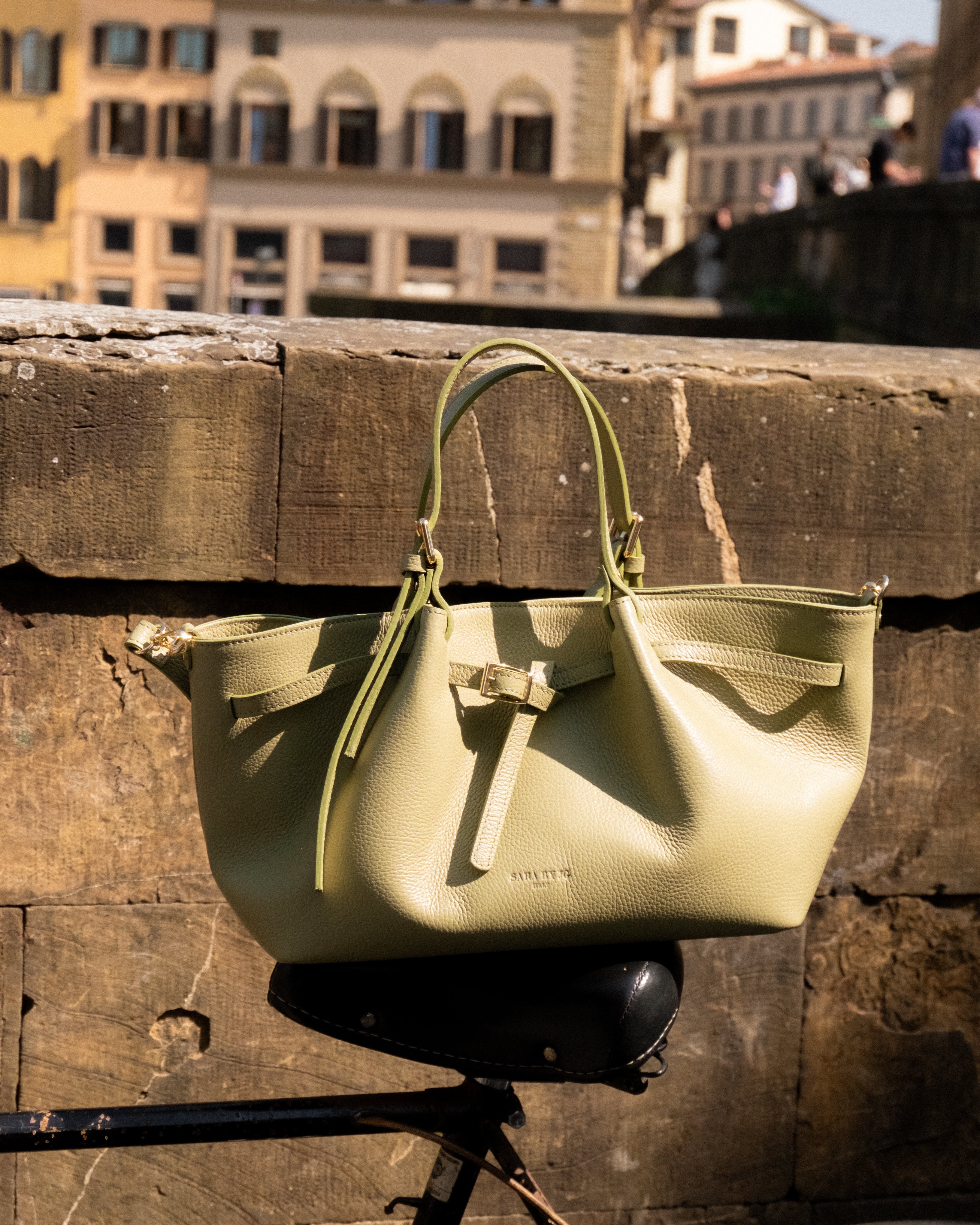 Italian leather handbag in olive green resting on a stone surface with a city backdrop.