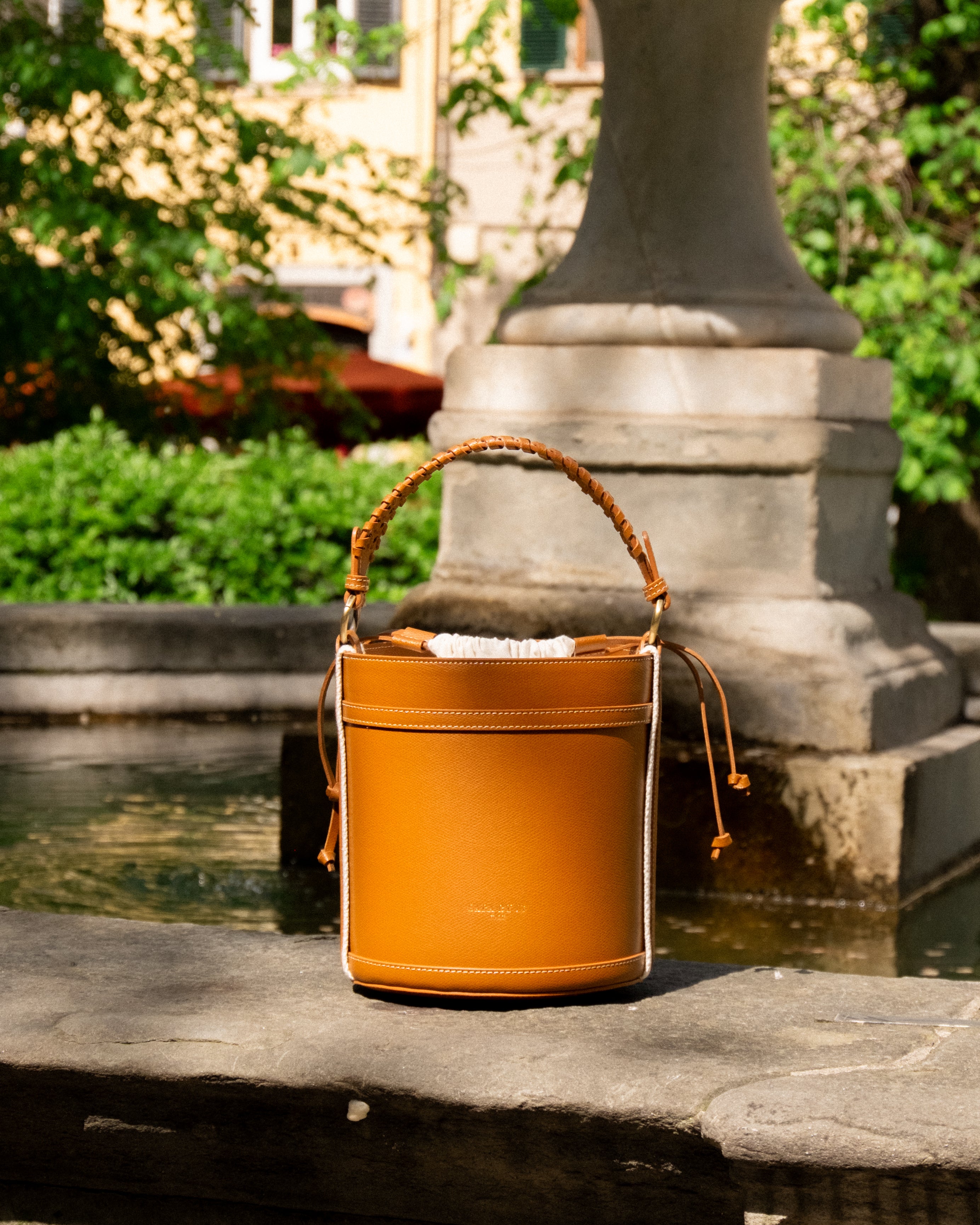 Modern minimalist bucket bag in orange with braided handle, displayed near a fountain surrounded by greenery.